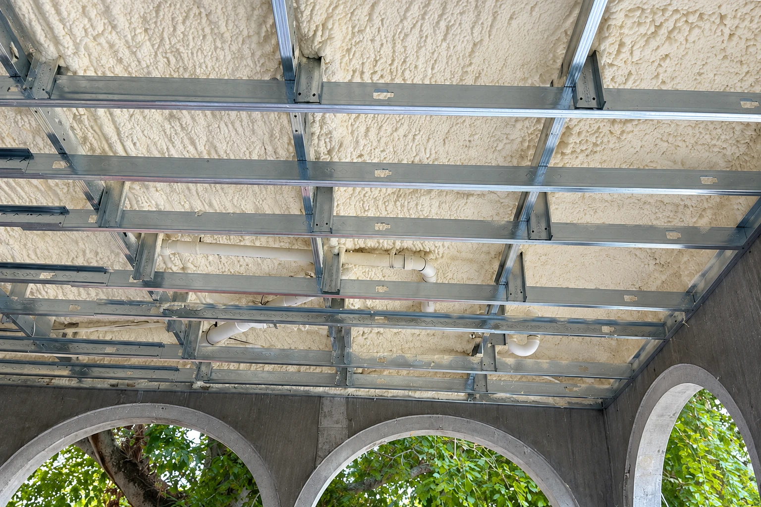 Spray foam coats the underside of a ceiling behind a metal framing grid, above concrete arches opening to greenery.