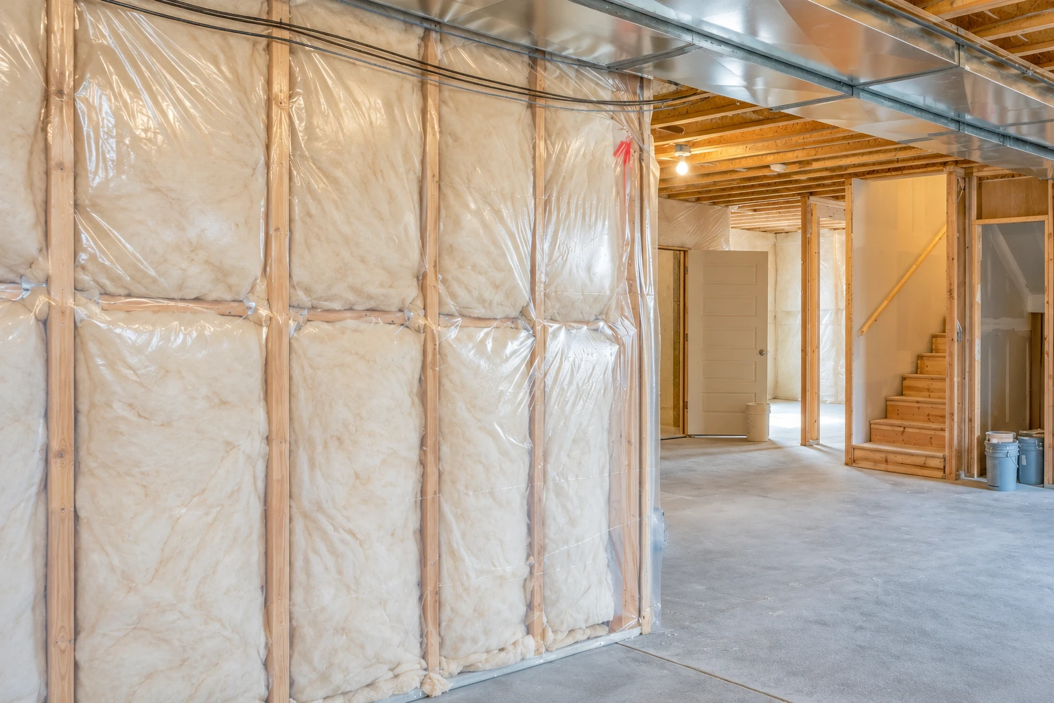 Fiberglass batts fill plastic-covered basement wall framing beside an open concrete-floored room with exposed ductwork and stairs.