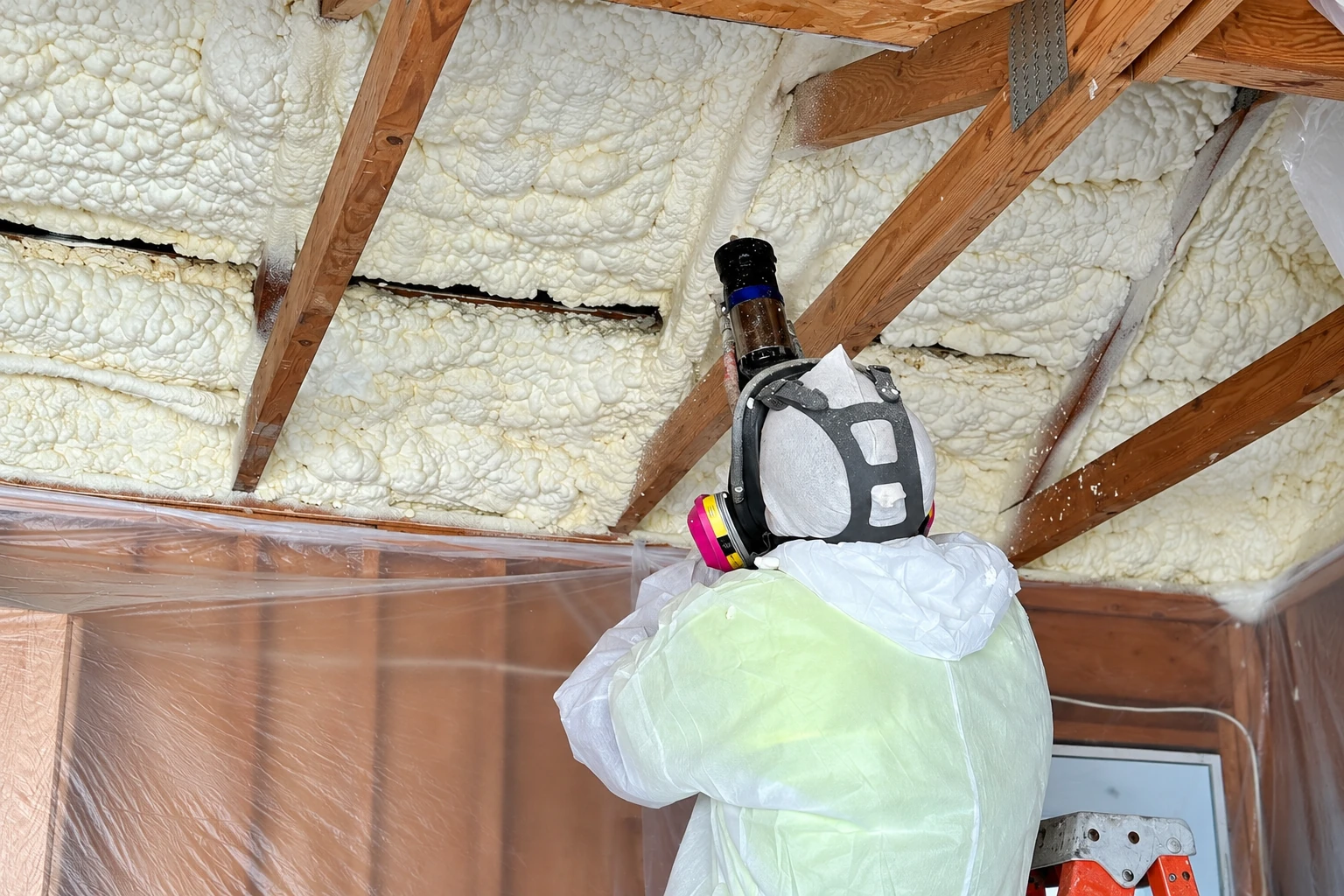 Worker in protective gear stands beneath roof rafters covered with thick white spray foam insulation.