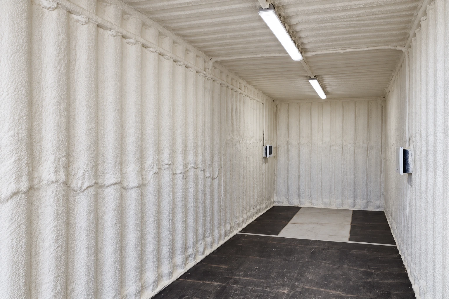 White spray foam coats the walls and ceiling of a long, empty commercial room with fluorescent lights.