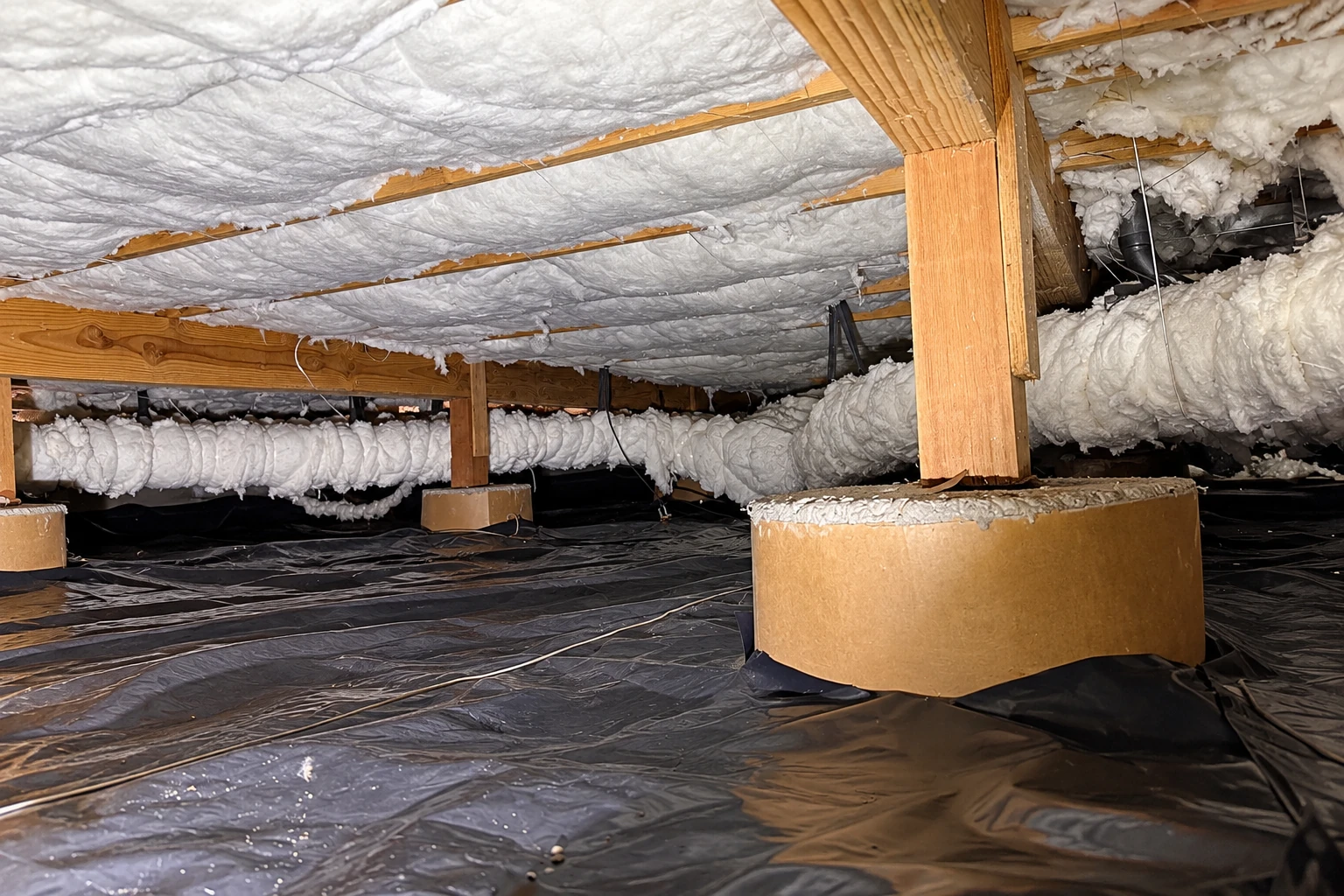 White insulation covers the crawl space ceiling and ducts above a black ground vapor barrier and concrete supports.